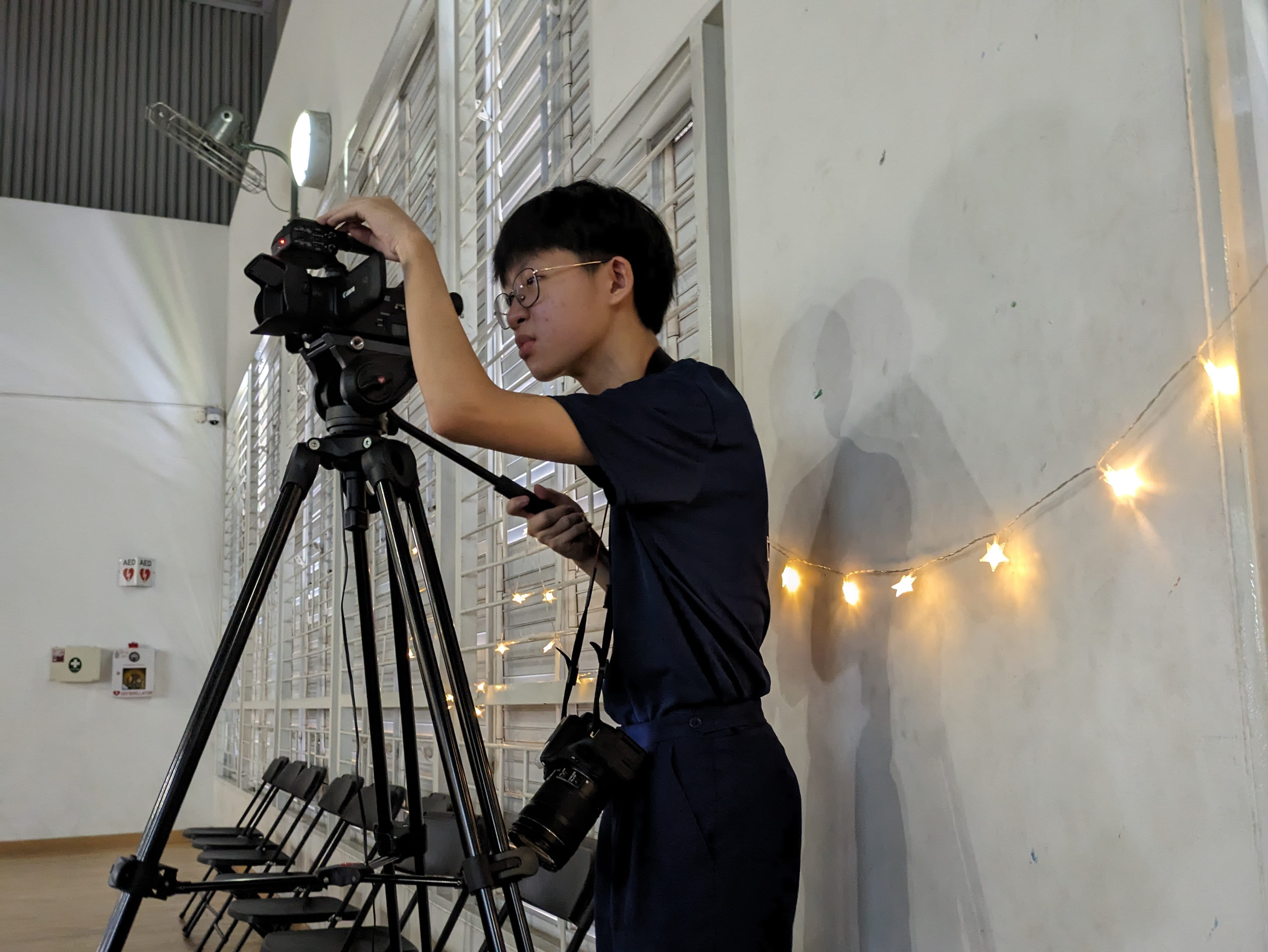 Student adjusting a Canon camera on a tripod indoors, with string lights on a white wall nearby.